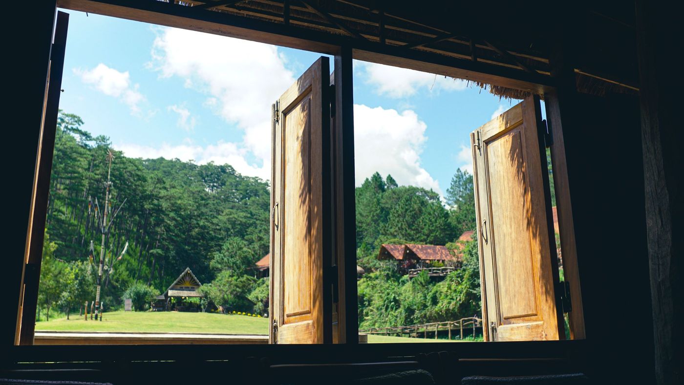 Open wooden window shutters looking out on green trees and a bright blue sky
